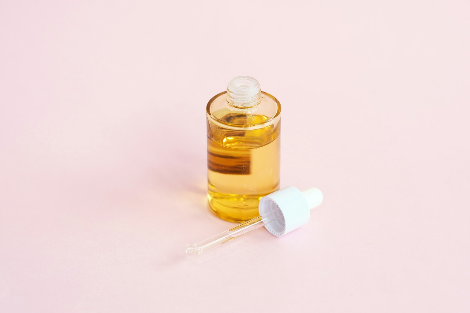 A single unlabeled glass bottle of facial oil with its dropper resting beside it on a soft pink surface.