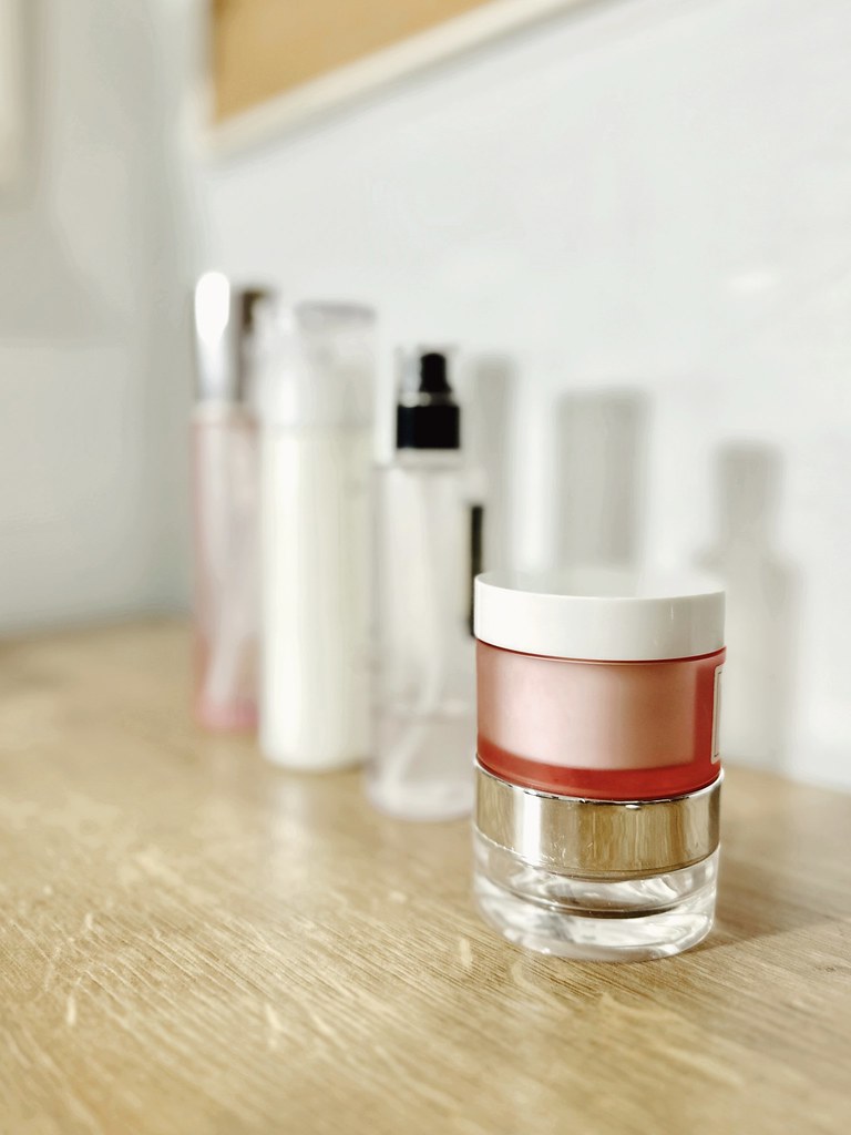 A rose-tinted cream jar in front of a soft-focus row of skincare bottles on a wooden shelf.